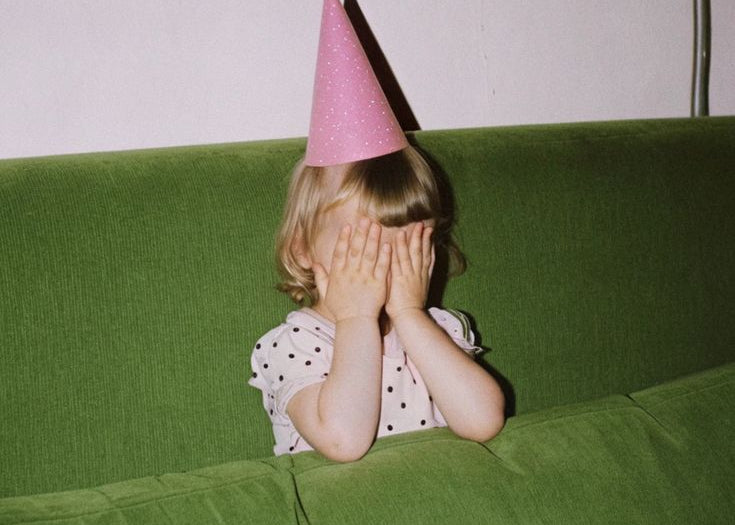 Child wearing a pink party hat sitting on a green couch with a plain background